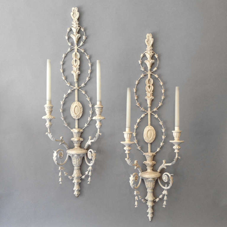 A pair of large scale carved and painted wall lights in the Adam taste, the back of each carved with intertwined husks above a bracket with two scrolling candle arms, one having beaded socles, the other with gadrooned socles.