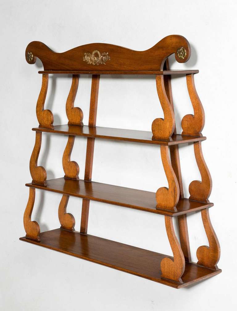 A pretty Regency period mahogany hanging shelves, the unusual scrolling uprights supporting four shelves, the pediment with an original gilt bronze applique.