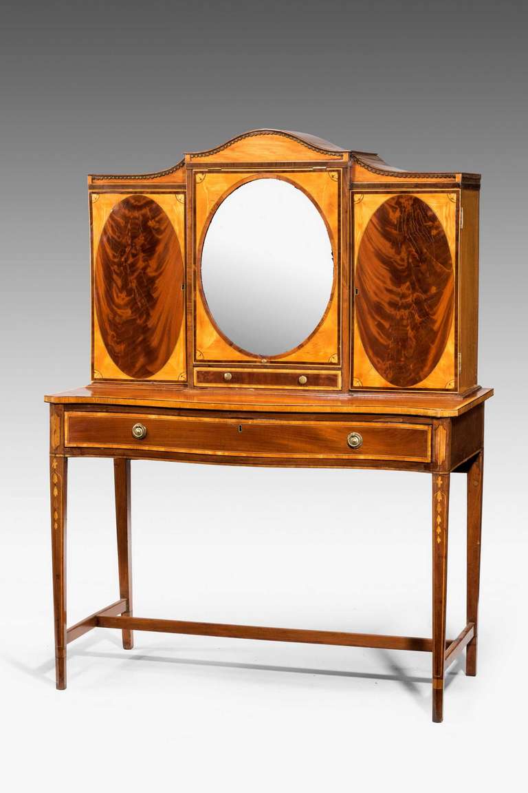 A George III period Irish mahogany dressing table, the central cabinet with an oval mirror flanked by beautifully flamed mahogany oval panels with a slender drawer to the centre section, the slender supports with hair bell inlay, the whole cross