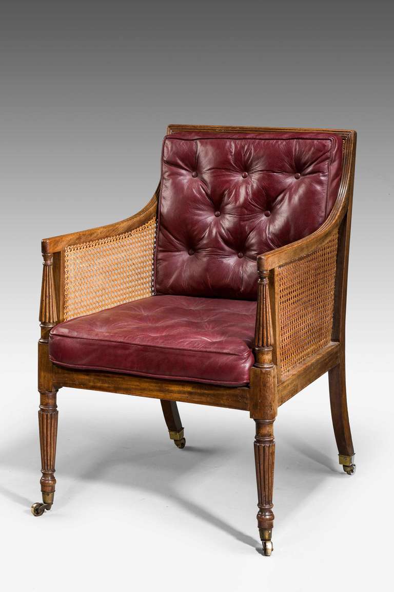 A Regency period mahogany bergere armchair, the uprights with fluted sections, the armrests with reeded carving. 

A bergère is an enclosed upholstered French armchair (fauteuil) with an upholstered back and armrests on upholstered frames. The