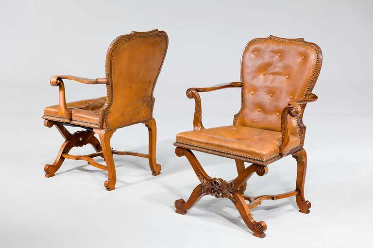 A pair of English mahogany Open Armchairs of George II style, early 20th century,each with a cartouch-shaped padded back and seats covered in buttoned close-nailed brown leather, the back and seat edged with ribbon and rosette, the scroll arms with