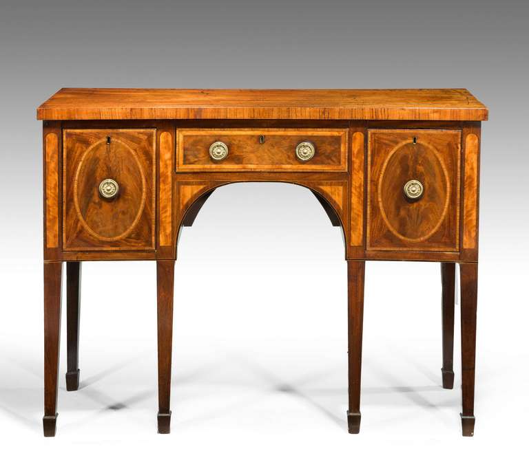 An unusual late 18th century concave Sheraton period mahogany sideboard with broad satinwood crossbanding to the top edge and the main carcass, tapering square section supports terminating in spade feet.

