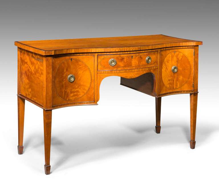 A late 18th century serpentine satinwood sideboard with a drawer to the left and a cupboard door to the right, cross banded top in rosewood standing on square tapering supports terminating in spade feet.


