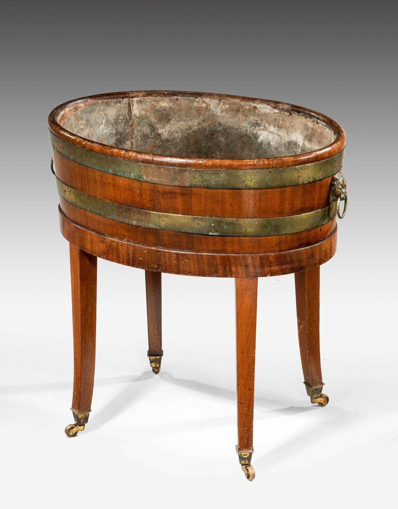 A Late 19th Century oval mahogany planter or champagne cooler. Bound with brass, of coopered construction on very slightly flared, tapering supports ending in the original shoes and casters.