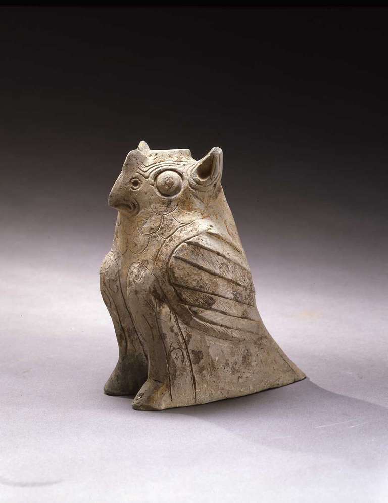 The grey pottery vessel modeled in the shape of a seated owl with pointed ears flanking the circular opening at top, a small hooked beak beneath a high bridge on the nose and two large, wide-set eyes. Carved overall with bold markings to stimulate