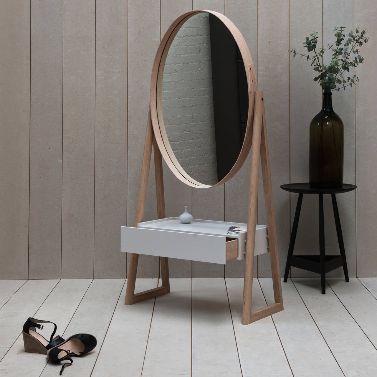 A full-length pivoting elliptical mirror with a shaker-style joint and brass rivet detail at one side held in an A-frame with a lacquered drawer box below.