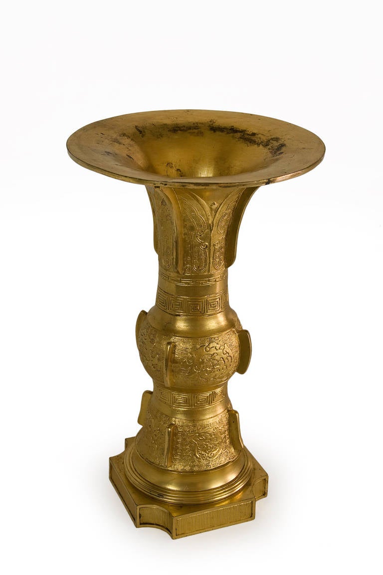 Chinese bronze GU form vase in gilded bronze. The upper flaring section with leaf-shaped panels, the central and lower sections with taotie masks and geometric decoration, all divided by shallow flanges. At the bottom a quadrangular base with curved