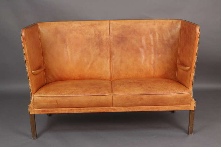 Freestanding high back two seater sofa with integrated armrests. Dark stained nutwood legs. Sides, back and seat in beautifully patinated natural leather fitted with brass nails. Made in 1943 by cabinetmakers Frits Henningsen. Acquired directly by