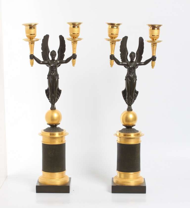 Both consisting of the patinated bronze goddess carrying a candle holder in both hands, standing on a sphere on a caneled cylindrical base on a square plinth.