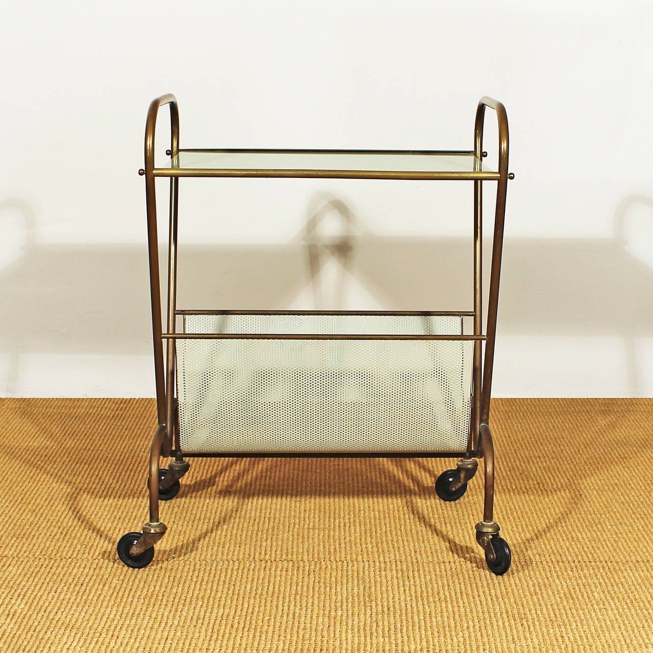 Bar cart and magazine rack, polished brass, white lacquered perforated sheet metal or rigitule, original wheels, coloured glass on top (minor losses).
In the style of Mathieu Matégot.
France circa 1950