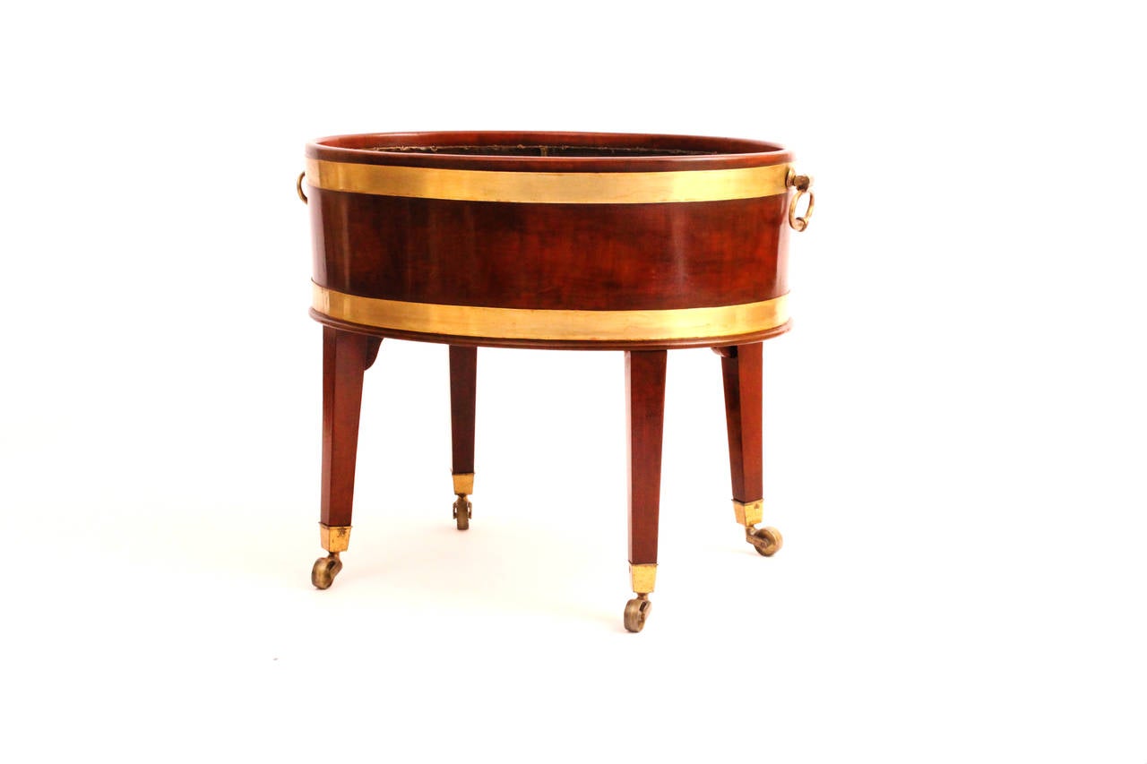 An oval mahogany cooler with brass bound and four vertical feet on castors. 
Two side brass handles.