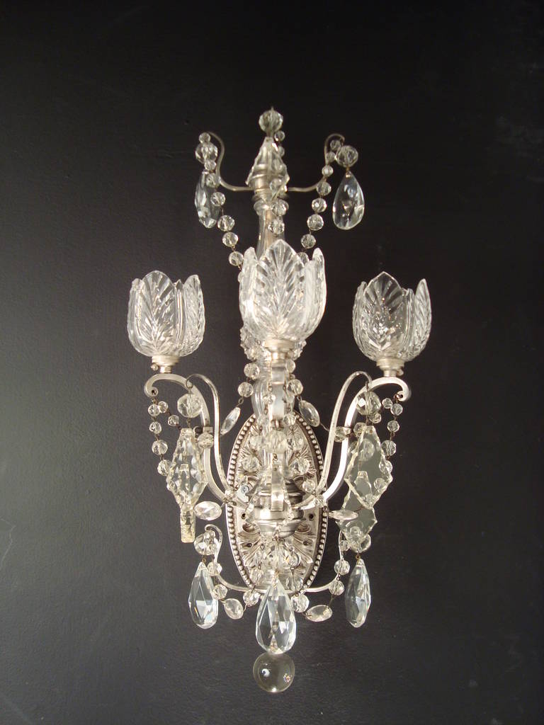A set of four Edwardian silvered bronze, glass and crystal three-light wall lights, by Faraday & Sons, The glass stems, issuing three scrolled bronze branches, with cut glass shades, the silvered bronze back plates of acanthus leaves design stamped