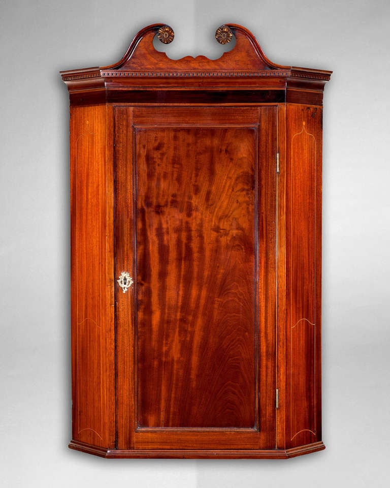 A particularly good and well detailed George III period mahogany and inlaid hanging corner cabinet surmounted by a scrolled pediment with carved gilt paterae above a dentil cornice and a contrasting inlaid frieze, the panelled door opening onto