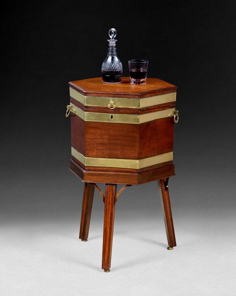 A very good George III mahogany and brass bound hexagonal wine cooler on stand, the lidded top opening to reveal a lead lined interior, the body retaining the original brass carrying handles and the whole raised on a conforming three legged stand,