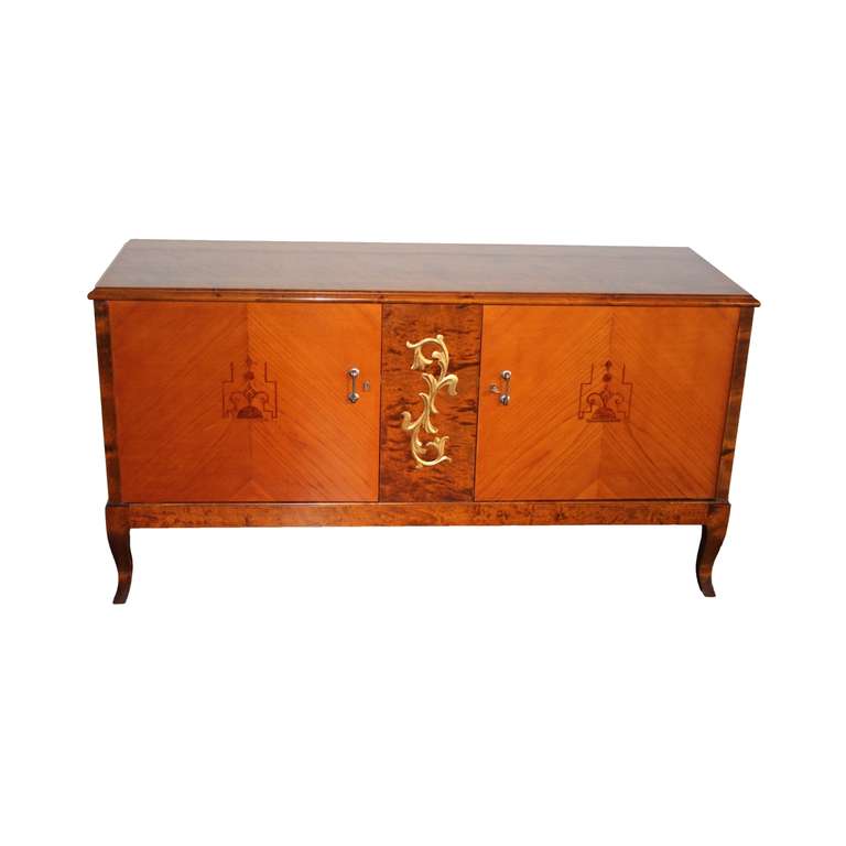 Flamboyant low server in flame birch raised on short cabriole legs. Two doors in elm decorated with geometric inlays in walnut, palisander, elm root and birch. Middle panel in flame birch adorned with a gilded acanthus leaf carving. Chromium plated