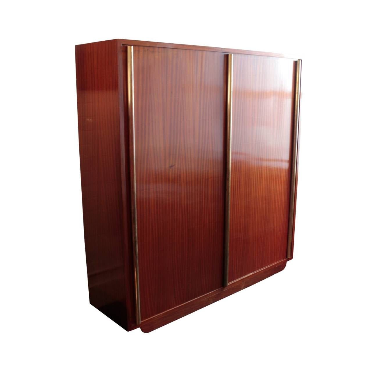 Large armoire in two sections having a pair of sliding doors with three full height cylinder shaped brass handles; in African ribbon mahogany, decorated throughout with Sornay patented miniature brass nail heads. Left interior section with hanger