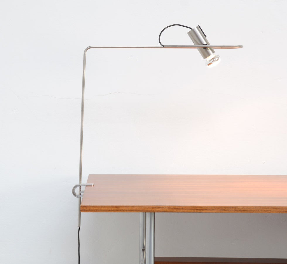 This minimal desk lamp is a design of the 1970s. It is a great and nice detailed lamp made of brushed stainless steel. The black bakelite switch is integrated in the cylindrical shade. 
This desk lamp is adjustable in height and the cylindrical