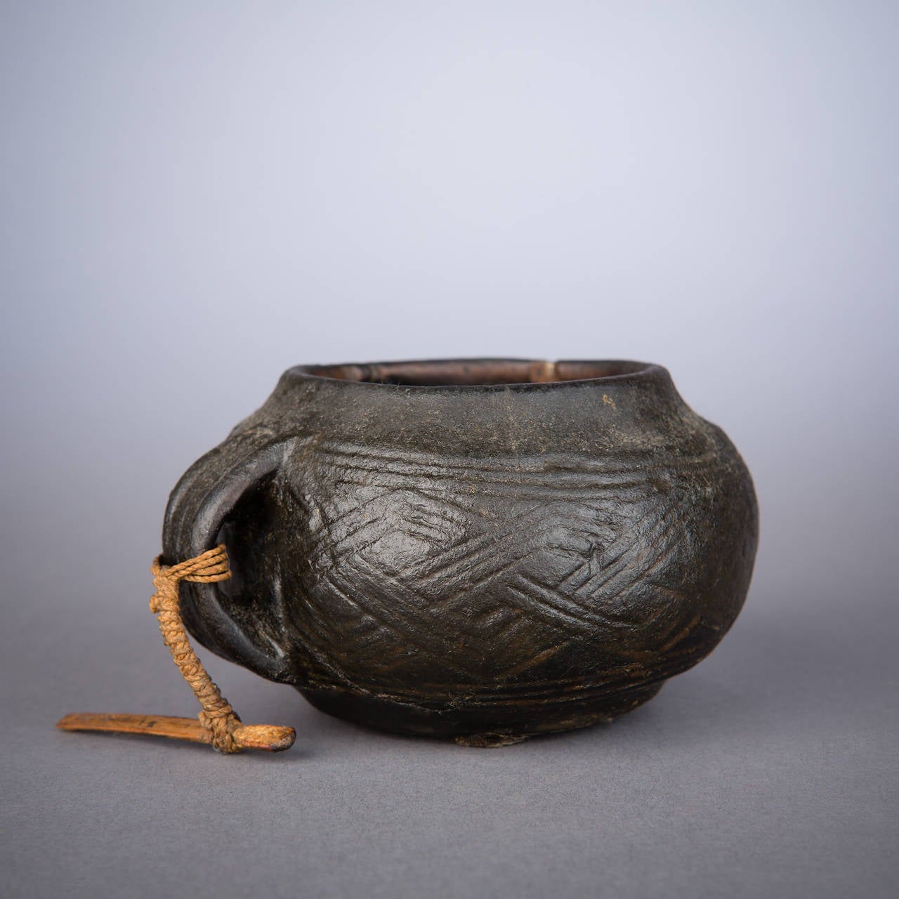 A very early Kuba cup heavily saturated with palm oil. Traditional Kuba geometric designs weave in faint relief around the cup's circumference. A fiber suspension cord remains attached to the handle. In good condition and old vertical split next to