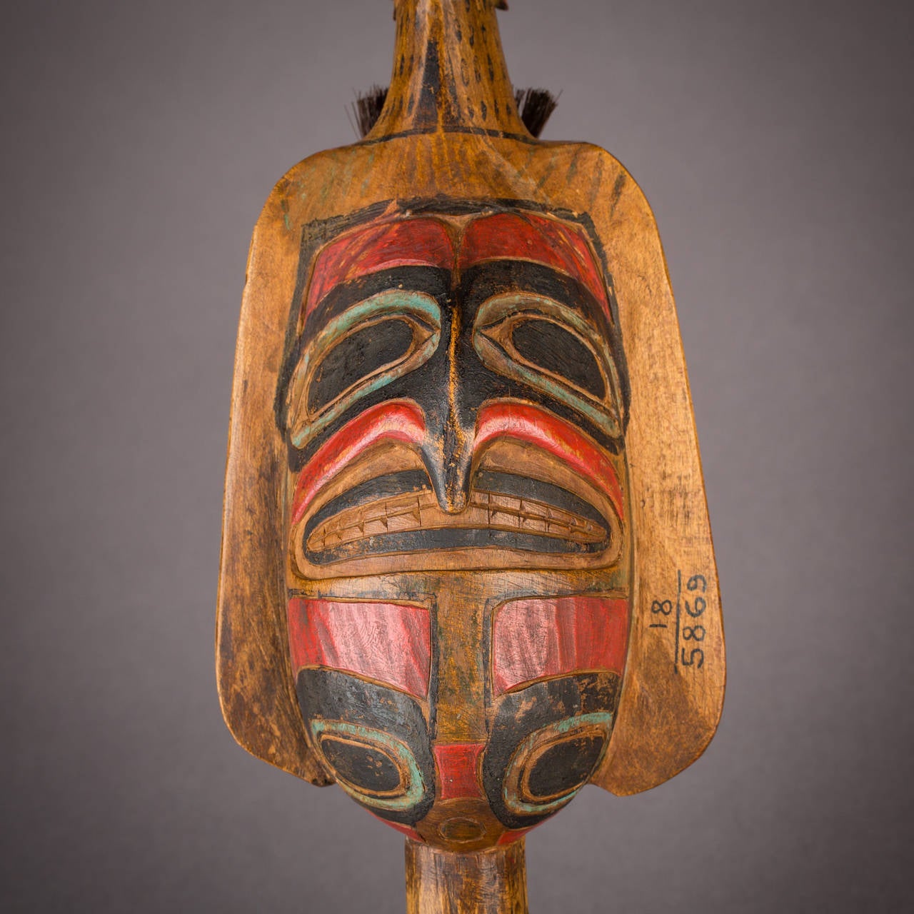 This Classic Bella Coola shaman's ceremonial rattle depicts a man reclining atop a large raven, his tongue held by a second raven head emerging from the primary bird's tail region. A stylized totemic face, possibly representing a hawk, is carved on
