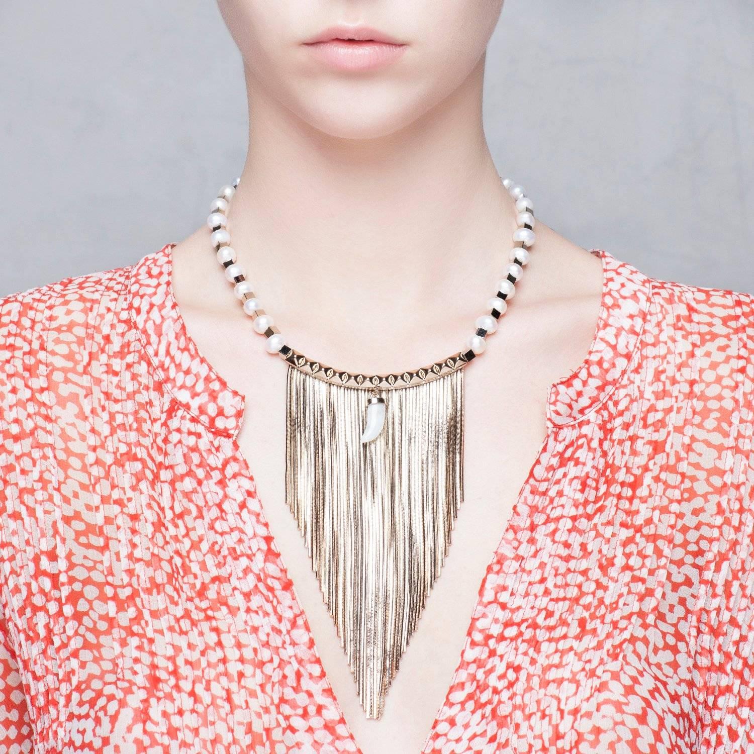 Bringing bohemian elegance to bib necklace, this fringed necklace features freshwater pearls alternated with gilded geometric brass beads. The piece is further enhanced by a studded central part with a jade horn shaped charm for an extra dose of
