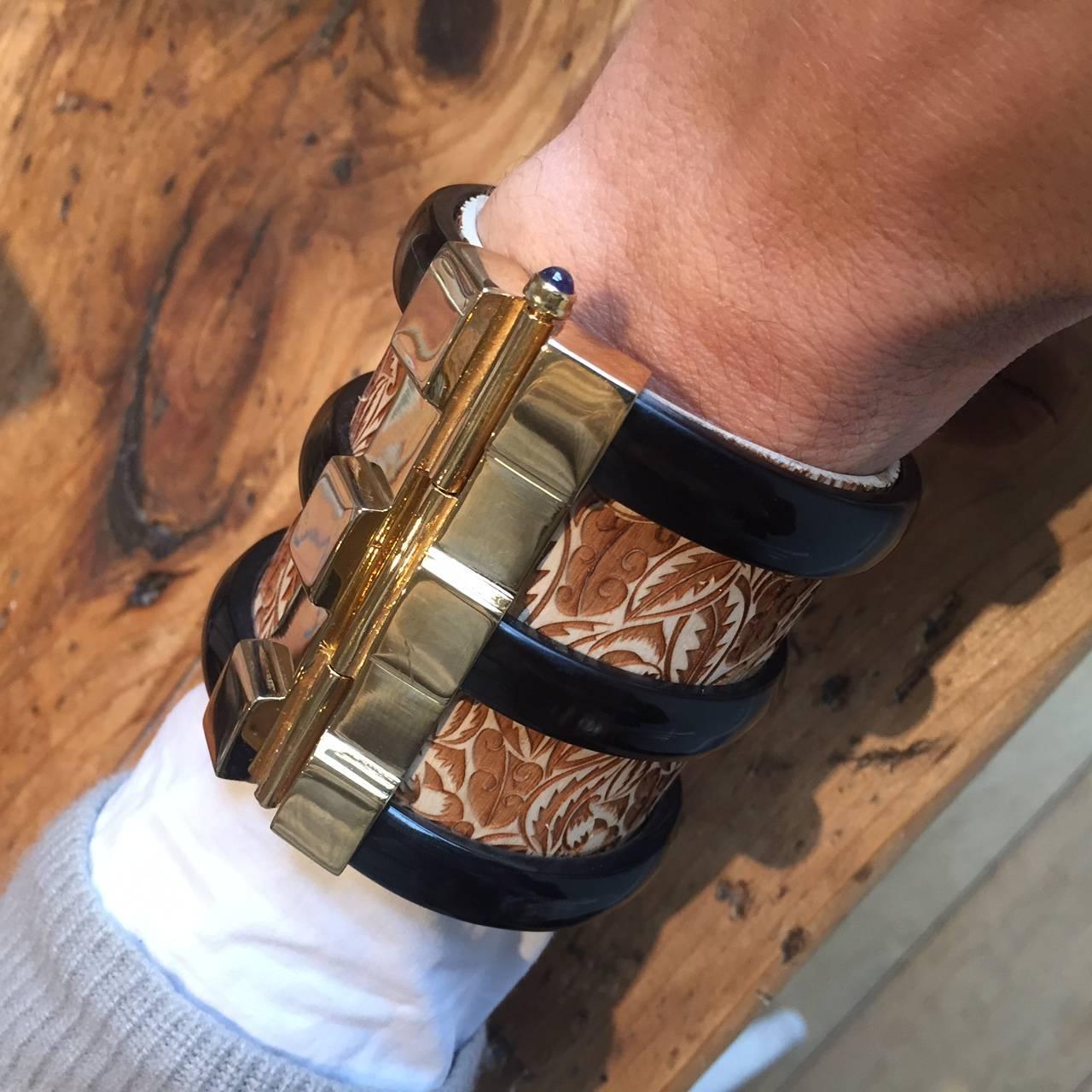 Cuff bracelet crafted from engraved jacaranda wood and African cow horn. The customizable 18k gold plated pin-clasp is set with a choice of ruby, emerald or blue sapphire. Inspired by warrior style cuffs worn by former Vogue editor Diana Vreeland.