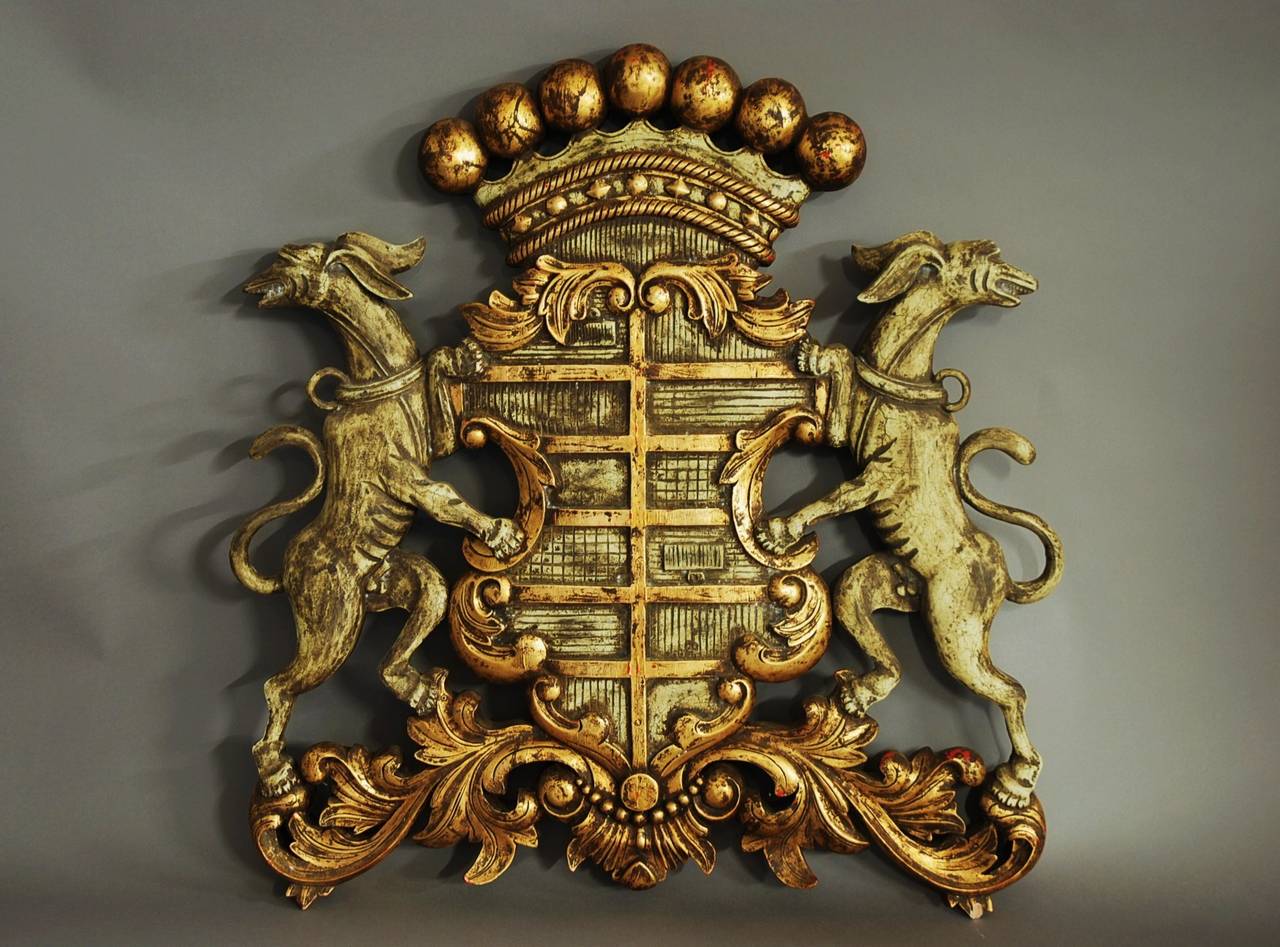 A highly decorative gilded & painted carved wooden coat of arms.

This coat of arms/armorial consists of a gilded crown to the top with a shield below finished with a pale green paint and gilding.

To each side of the shield there are mythical