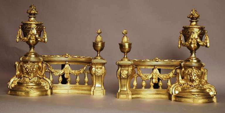 A decorative pair of late 19th c French Louis XVI style ormolu (gilt brass) chenets/fire dogs. 

Each chenet has a large Classical urn with a flame and decorated with swags. 
The urn is supported by a column with a lions head to the