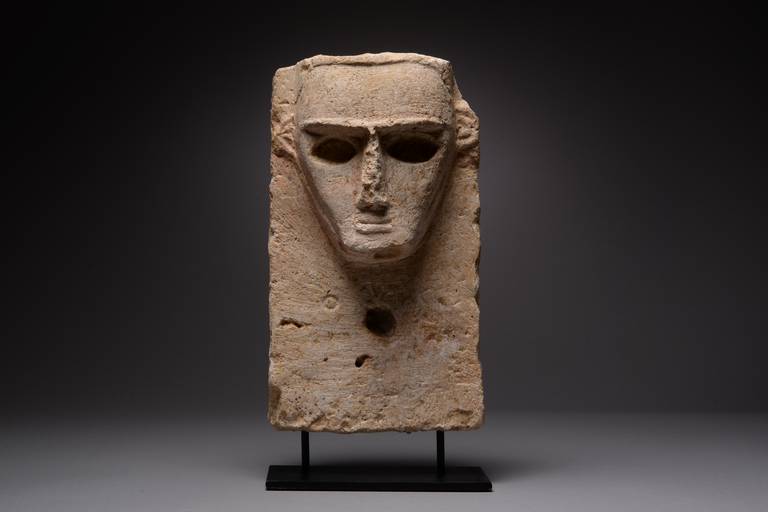 An ancient south Arabian limestone pillar stele, dating to the 3rd–1st century BC.

The head sculpted in relief with robust features; prominent ridge, long nose, almond-shaped hollow eyes and a terse mouth. A Sabaean inscription runs beneath,