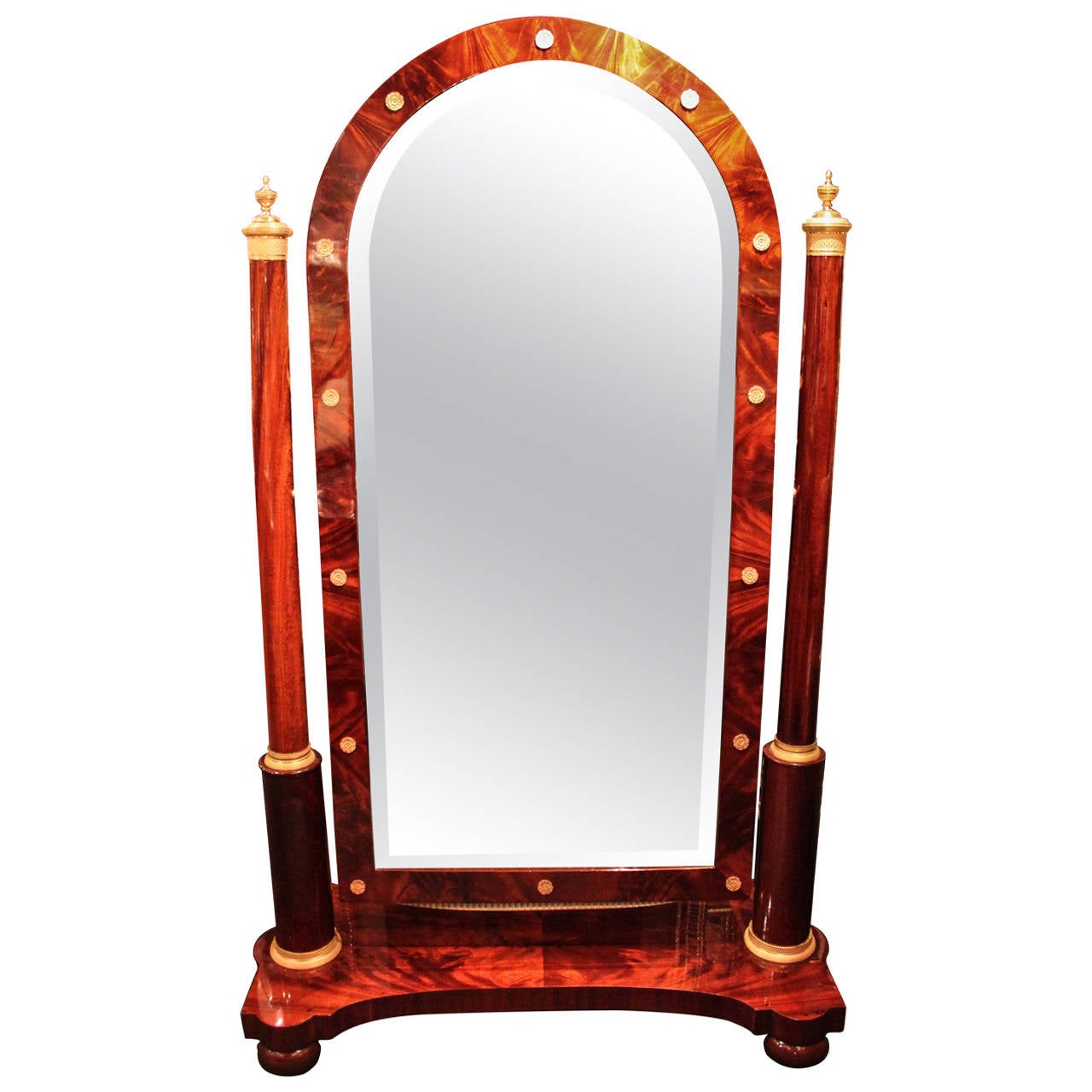 A fantastic example of a mid-19th century cheval mirror in the First Empire style. Two round columns surmounted by covered, gilded bronze cups support the curved and beveled mirror, decorated with gilt rosettes. Rich mahogany veneers with a deep