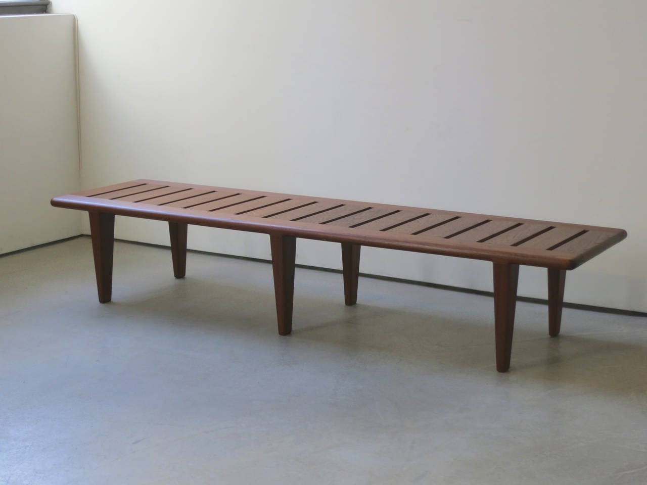 Beautifully proportioned low bench in solid teak with six tapered legs. Designed by Hans Wegner and made in the 1950s by cabinetmaker Johannes Hansen.