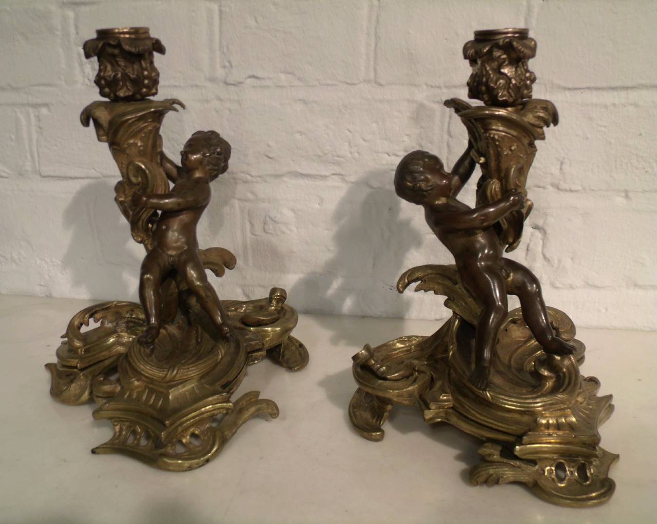 This very attractive pair of French bronze and gilt bronze candelabras feature a pair of poising cherubs on a very decorative floral centre stem and heavily scrolled base. The top half of the candelabras can be removed to be used for a single taper