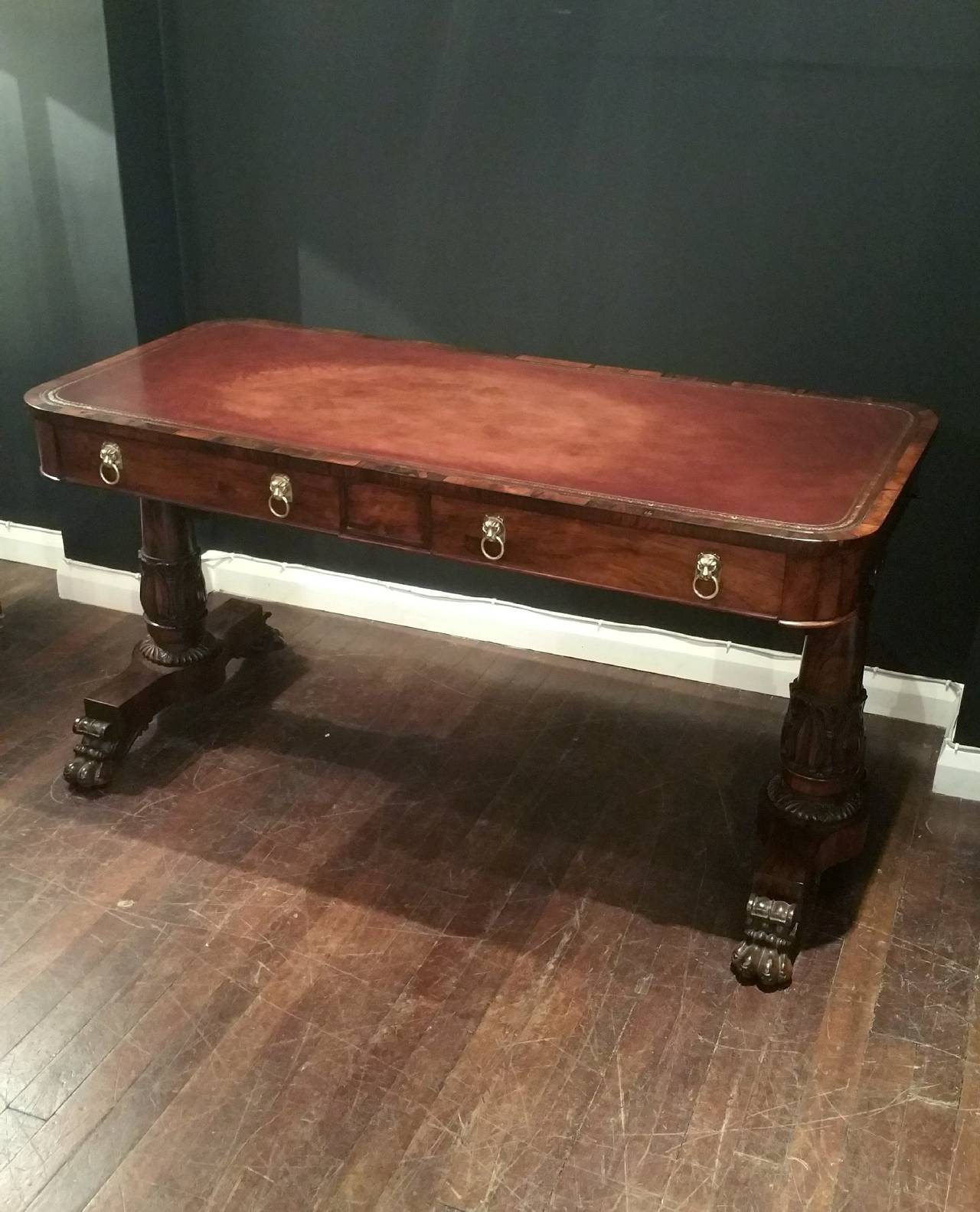 This superb and very handsome Regency Rosewood library table features a red brown leather tooled top with lion’s head and brass ring drawer pulls. The table stands on carved column supports and end with hairy paw feet on brass castors. The table