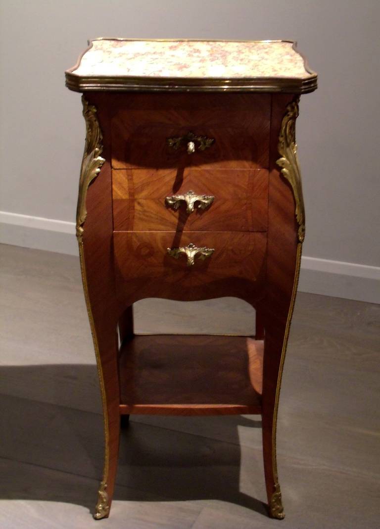 A superb pair of turn of the century marble topped Kingwood bedside tables with ormolu mounts and trim. The bombe shaped tables feature three drawers with ornate ormolu drawer pulls and a lower bottom shelf with a handsome parquetry design. The
