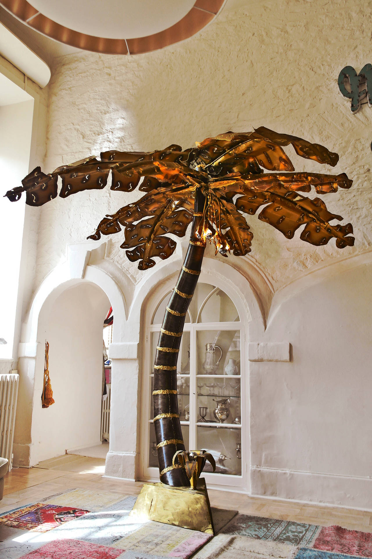 Monumental palm tree floor lamp in torch cut brass, brutalist style. Features illuminating pendent bulb design and second lamp at base.