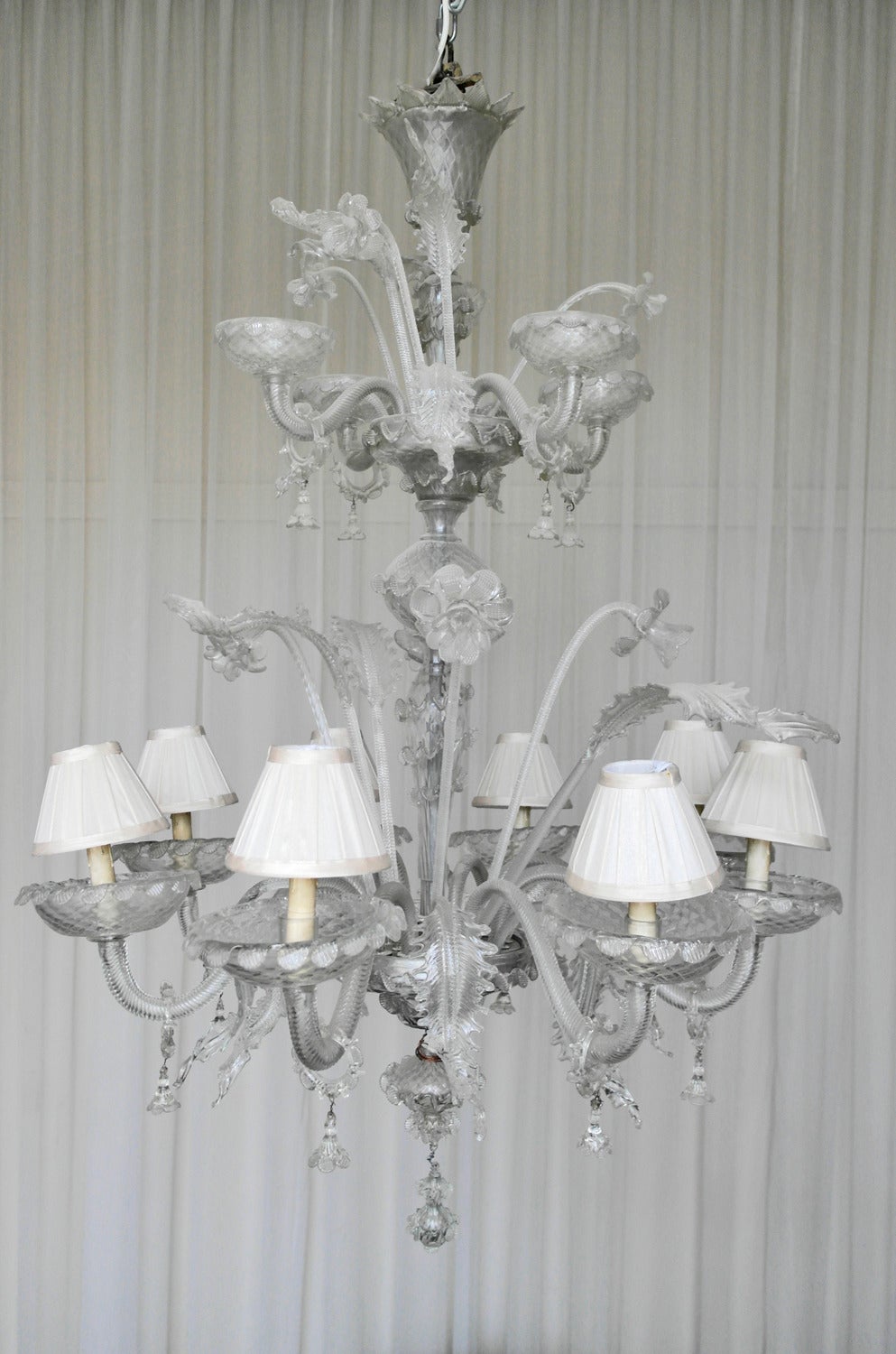 Eight arm, two tier chandelier in Murano glass with organic floral detailing. Each candle arm features a dripping-wax effect candlestick light with pale gold satin lampshades (estimated to be a later addition to the piece.) Ideally suited to grand