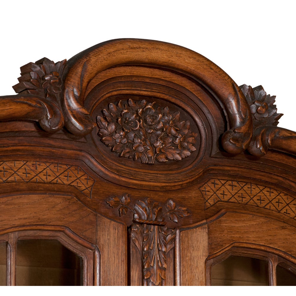 Two-part French country oak cabinet with arched and carved pediment and mullion glass doors in the top. Base has two drawers over carved panel doors. Hand-carved detail with the Classic French motif of flowers and leaves, circa 1900. Wear consistent