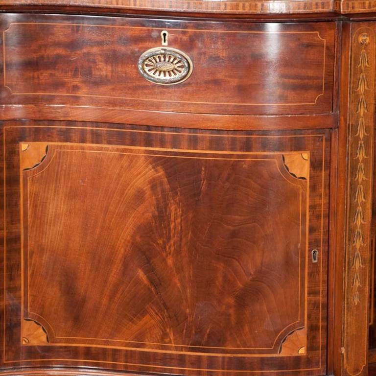 19th century English Sheraton solid mahogany sideboard with bowed front, satinwood quarter fan and bell flower inlay, brass gallery and tapered legs. It features three drawers and four doors with ample storage for silver, China and linens.