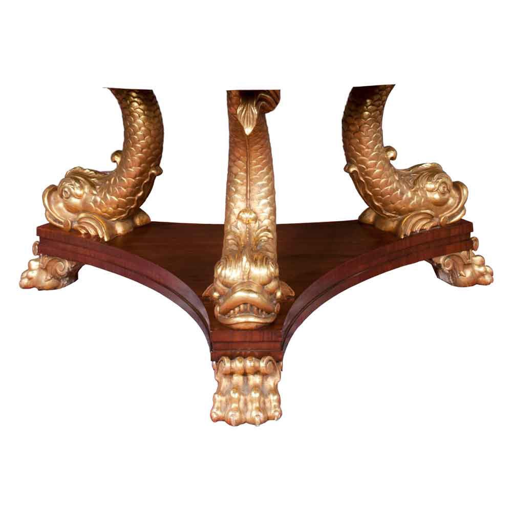 English Regency style library table with cross-banded rosewood top. The base features three fully hand carved gilded dolphins, a triangular shaped platform base and three gilded claw feet.