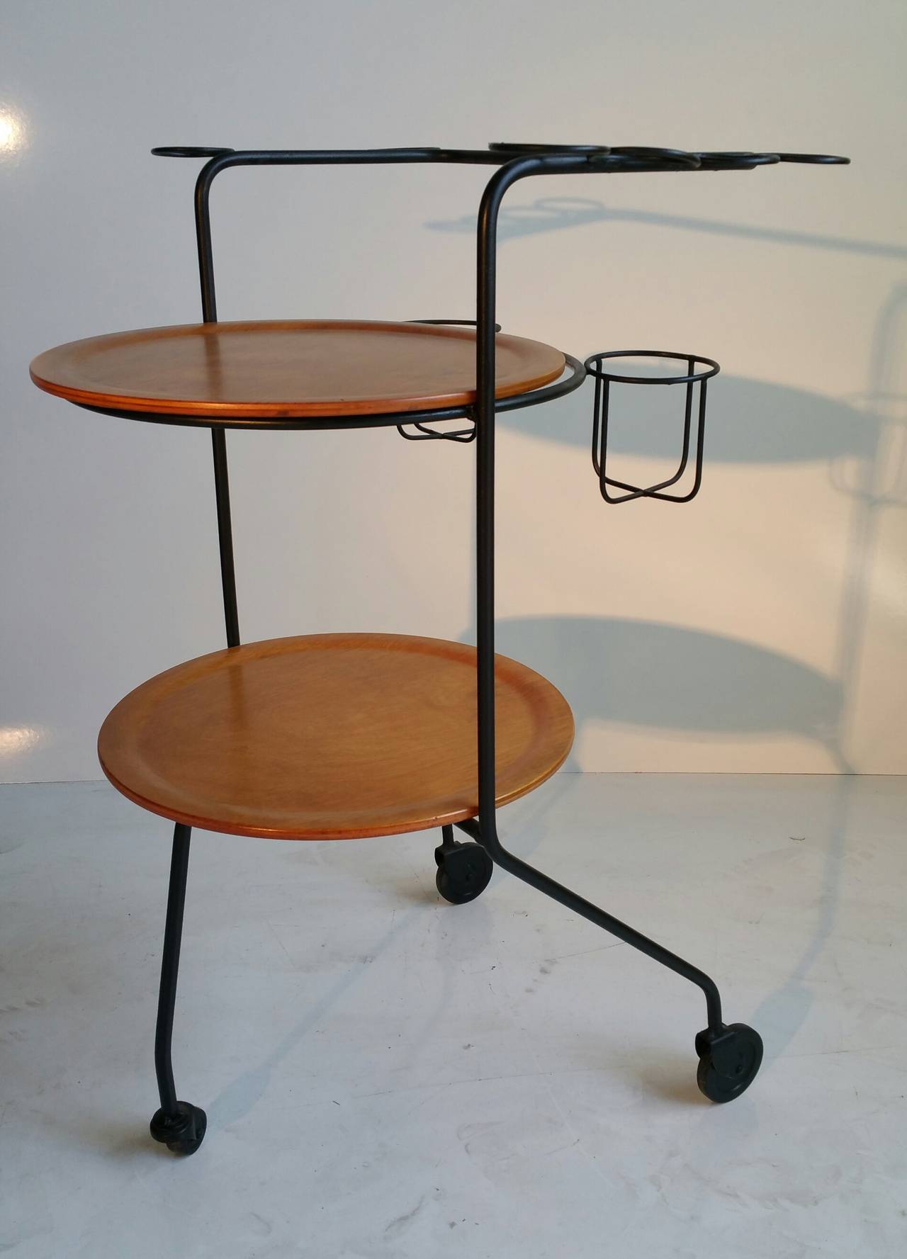 Two-tier rolling bar cart in tubular black metal with eight glass holders, two bottle holders, and wood shelves. Designed by Tony Paul, USA, 1950s.