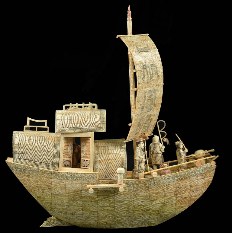 A large Chinese bone model of a leisure fish boat finely incised all-over,
The prow of the boat is occupied by a crew of three male figures holding a net, a fishing rod and a staff beneath a tall mast with inscribed banner,
Canopy or cabins