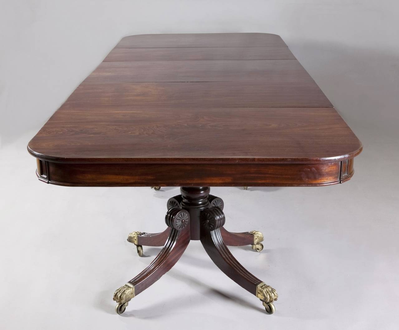 A superb George IV mahogany three-pillar dining table. The exquisitely figured mahogany top, resting on three turned four splay sabre leg carved pillars, with star mounts and large brass lions paw feet.