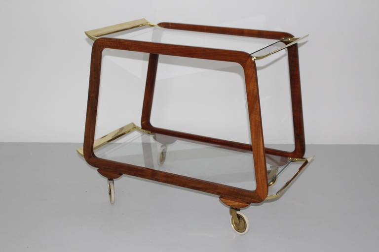 A mid century modern vintage brown walnut and brass bar cart, which was designed and executed in Austria 1960s. Also the bar cart features wheels for mobility. Two tiers were made of clear glass plates.
The materials of warm brown tone walnut and