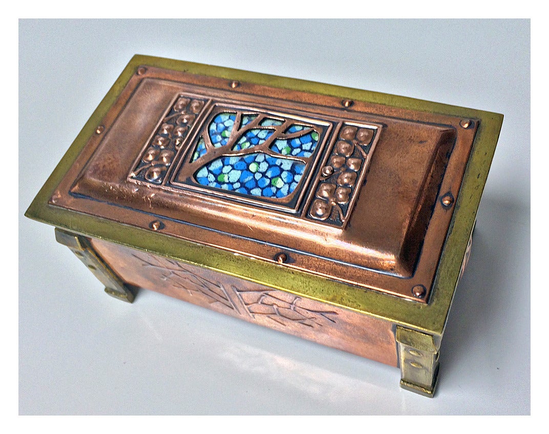 Arts and Crafts enamel, copper and brass box, circa 1900. The box of four turned rivet bracket brass cornices, the panelled copper body with stylised arts and crafts decoration, the cover with turquoise, white and green pebble design enamel