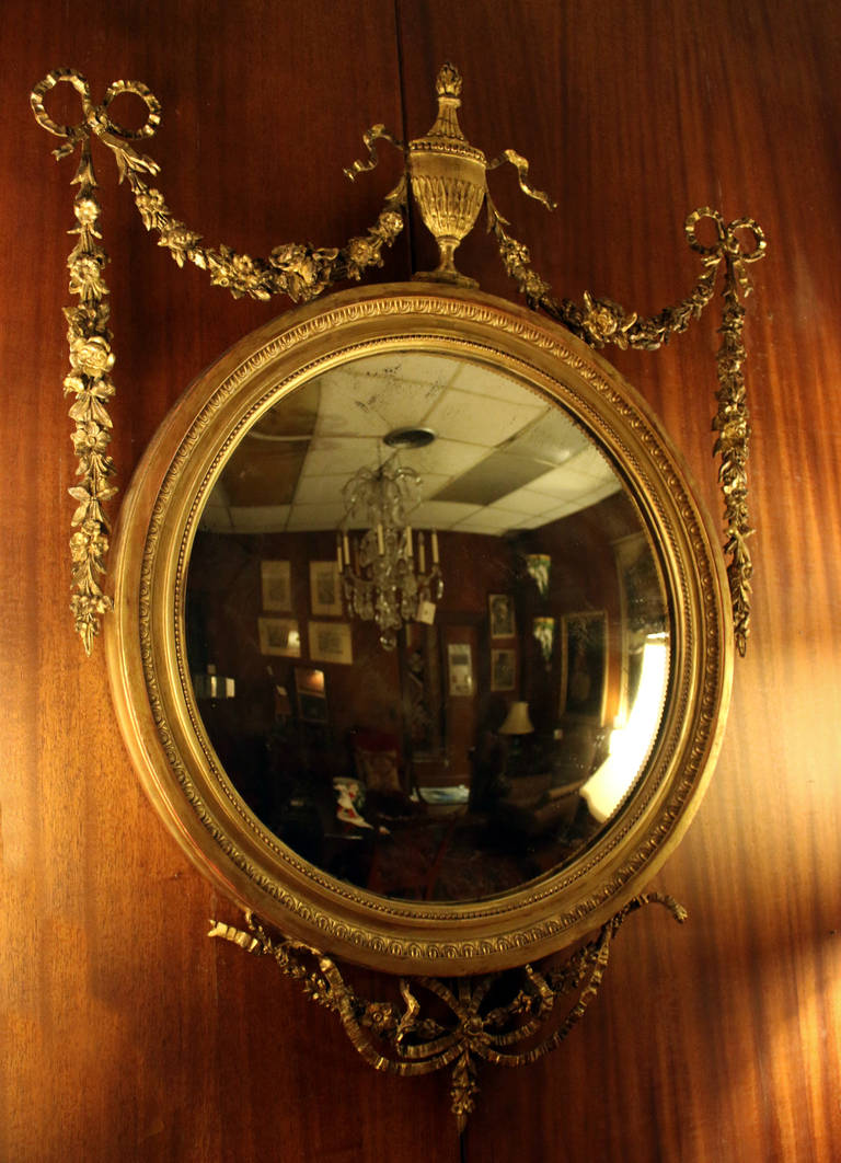 19th century convex mirror of the style of the Brothers Adam. English, constructed of wood with gold gilt. Features include delicate gessoed swags of flowers, ribbons, bows and a graceful flaming urn at the top. It has a slightly pronounced curve to