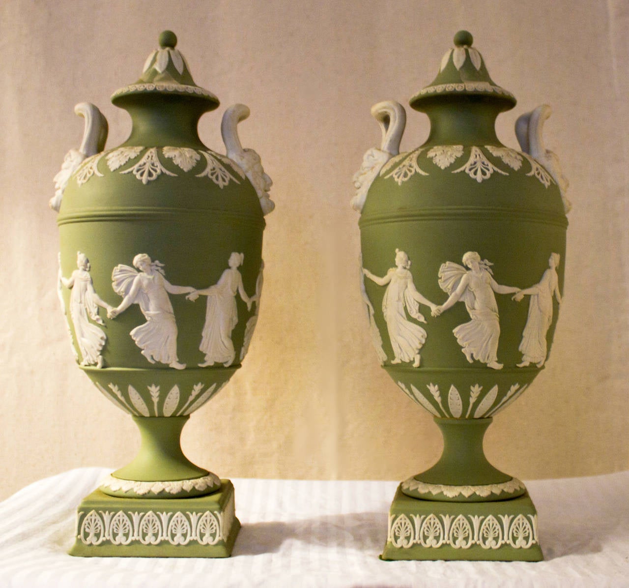 Pair of pale green Jasperware presentation lidded urns feature a white bas-relief ornamentation depicting Neoclassical Greek revival muses. The delicate handles on either side are molded over Bacchus heads and classical designs outline the lid and