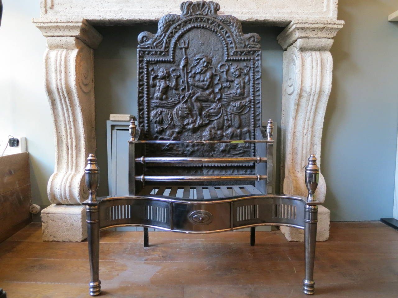 A late 19th century Georgian style fire grate in polished steel and cast iron. The decorative fireback of Poseidon, tapering legs surmounted by classical urn finials and serpentine fluted fret with oval patarae to centre.