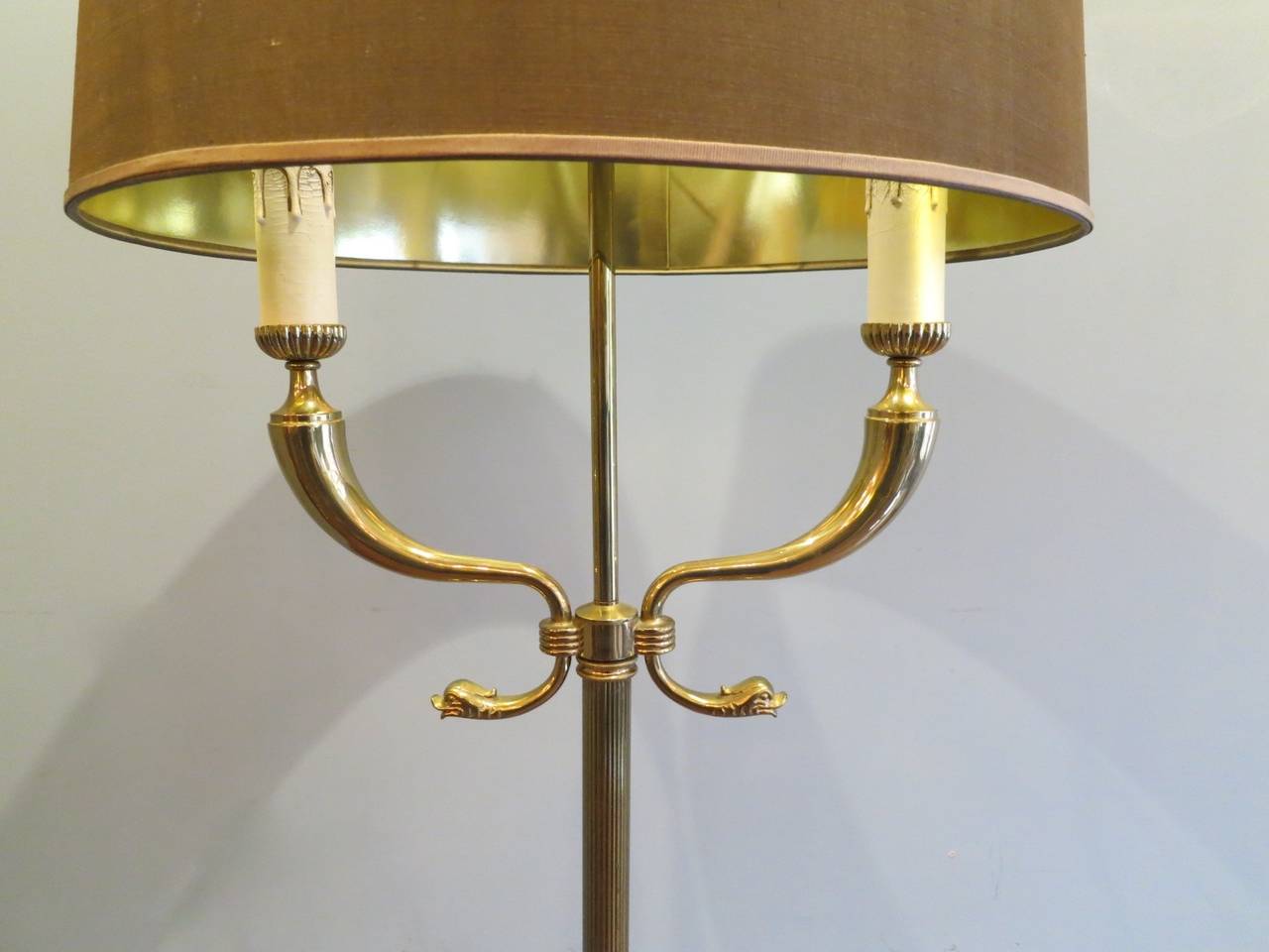 A large twin arm brass neoclassical style floor lamp, with Triton head arms brass base with inline switch and top fitting finial shade in bronze/gold color.