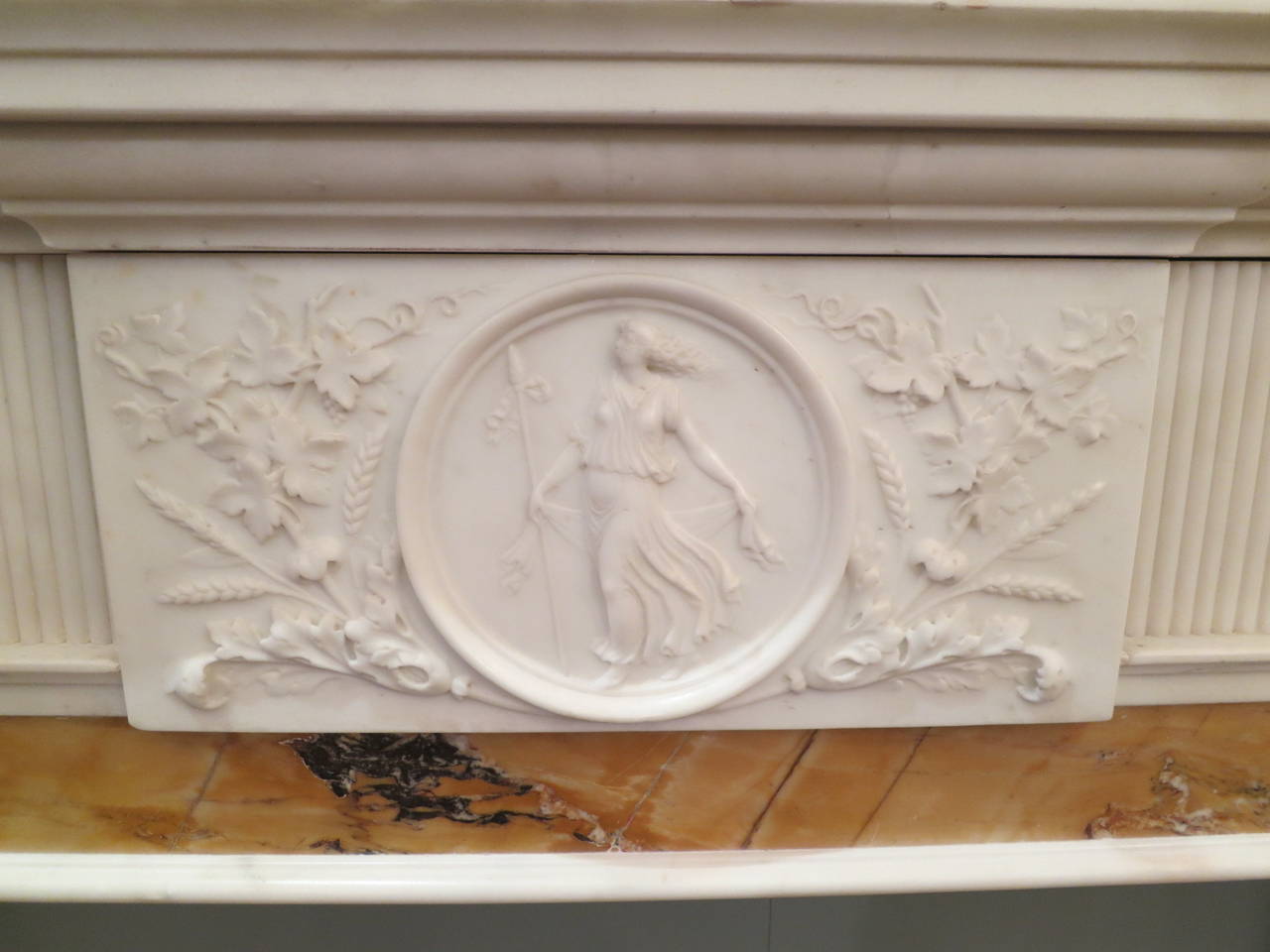 A statuary white and sienna inlaid marble fireplace in the neoclassical manner. Finely executed carved panels to jambs depicting the Arts, with classical figures playing instruments. There are also flutes, actor’s masks and pipes. The centre tablet