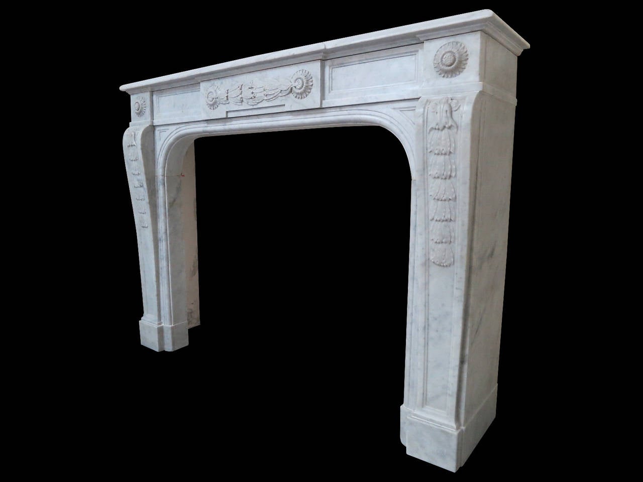 An unusual Louis XVI style fireplace in Carrara marble. The tapering console jambs decorated with carved ribbon tied bell drops and surmounted by corner blocks of circular floral carving. The Frieze with conforming carving to center plaque flanked