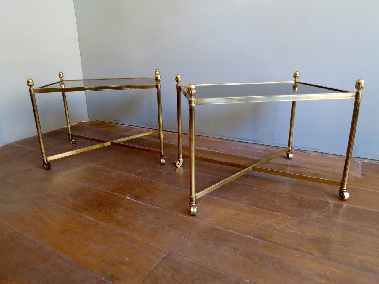 A large pair of side tables in brass with original smoked glass tops, X-stretcher supports, brass castors and brass acorn finials.